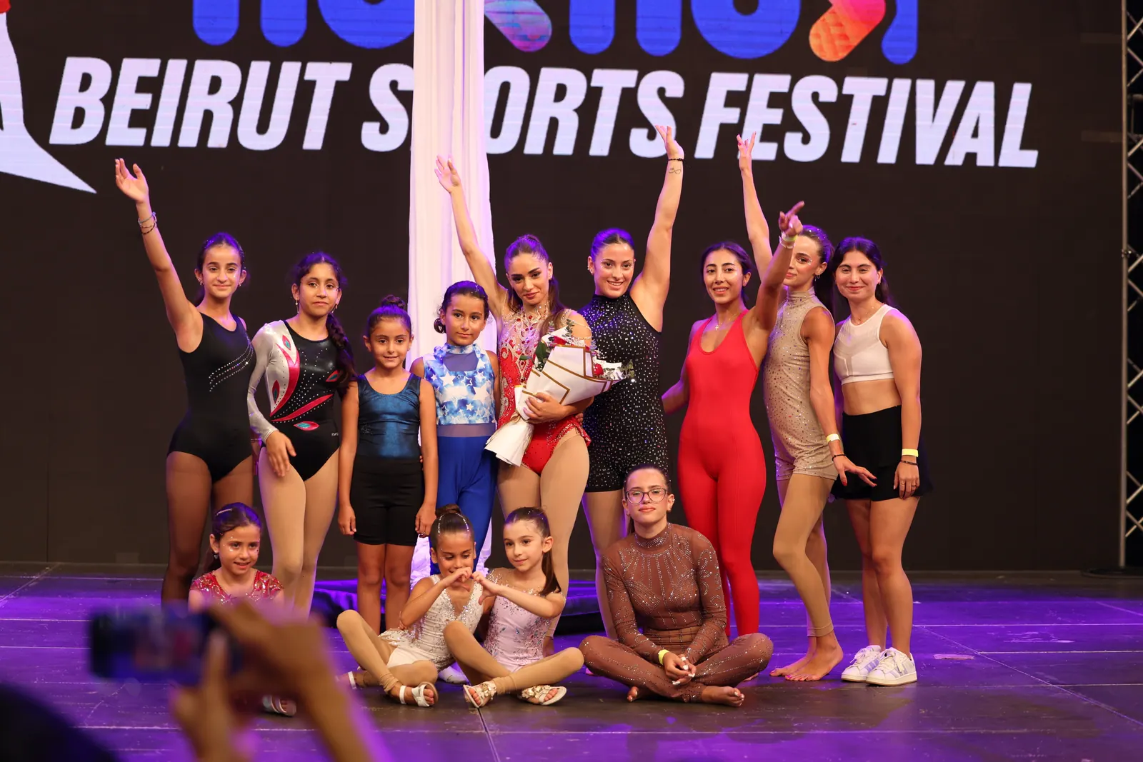 Students of Gaelle Matar Aerial Academy on stage together at the Beirut Sports Festival, from young children to adults, beside a white aerial silk.
